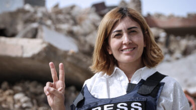 Lübnanlı gazeteci Amal Khalil üzerinde PRESS yazılı çelik yelekle enkaz önünde zafer işareti yaparken.