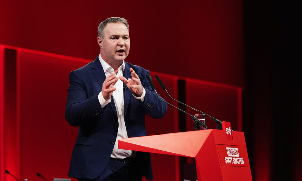 SPÖ lideri Andreas Babler, kırmızı bir arka plan önünde kürsüde hitap ederken elleriyle vurgulu bir hareket yapıyor.