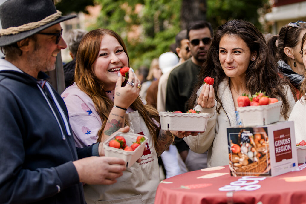 Festival alanında ellerinde taze çilek sepetleri tutan ve tadım yapan neşeli insanlar. Masada AMA Genuss Region logosu görünüyor.