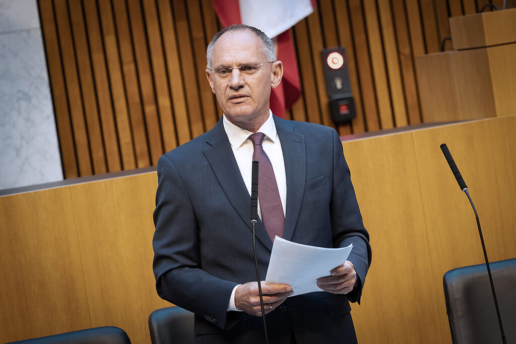 Avusturya Ulusal Meclisi (Nationalrat) oturumunda konuşma yapan bir milletvekili, arka planda meclis üyeleri ve Avusturya bayrağı.
