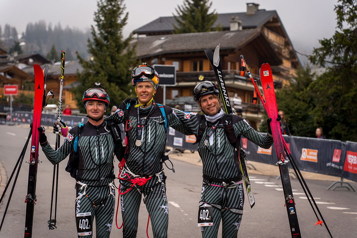 İsviçre Alpleri'nde Patrouille des Glaciers 2026 yarışını kazanan üç Avusturyalı Bundesheer askeri, kayak takımları, kaskları ve yarış numaralarıyla (402) podyum öncesi gülümseyerek poz veriyor.