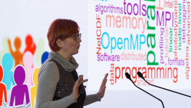 AI Factory Austria Öğrenme Merkezi Başkanı Claudia Blaas-Schenner, arkasında yapay zeka ve programlama terimlerinden oluşan bir kelime bulutu panosuyla sunum yaparken.