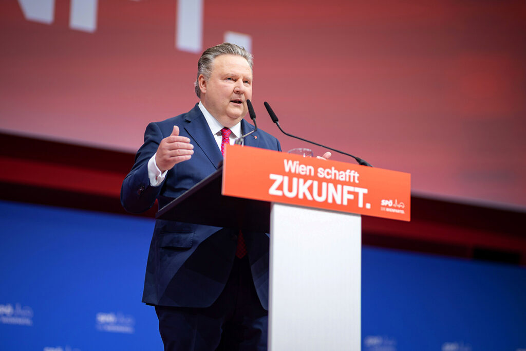 Viyana Belediye Başkanı Dr. Michael Ludwig, üzerinde 'Wien schafft ZUKUNFT' yazılı turuncu bir kürsüde konuşma yaparken.