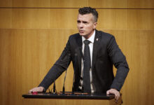 FPÖ Güvenlik Sözcüsü Gernot Darmann, Avusturya Parlamentosu kürsüsünde konuşma yaparken kararlı bir ifadeyle kürsüye tutunuyor.