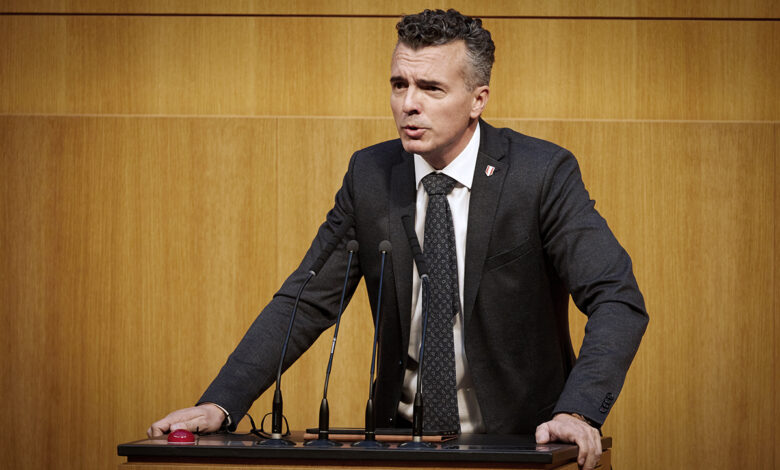 FPÖ Güvenlik Sözcüsü Gernot Darmann, Avusturya Parlamentosu kürsüsünde konuşma yaparken kararlı bir ifadeyle kürsüye tutunuyor.