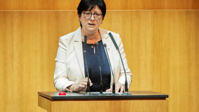 FPÖ Milletvekili Rosa Ecker, üzerinde siyah bir ceket ve kırmızı bir bluzla Avusturya Ulusal Meclisi (Nationalrat) kürsüsünde mikrofon başında konuşma yapıyor. Arka planda meclis salonunun ahşap detayları ve meclis başkanlığı kürsüsü görülüyor.