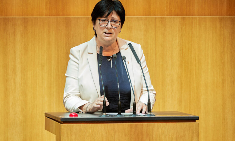 FPÖ Milletvekili Rosa Ecker, üzerinde siyah bir ceket ve kırmızı bir bluzla Avusturya Ulusal Meclisi (Nationalrat) kürsüsünde mikrofon başında konuşma yapıyor. Arka planda meclis salonunun ahşap detayları ve meclis başkanlığı kürsüsü görülüyor.