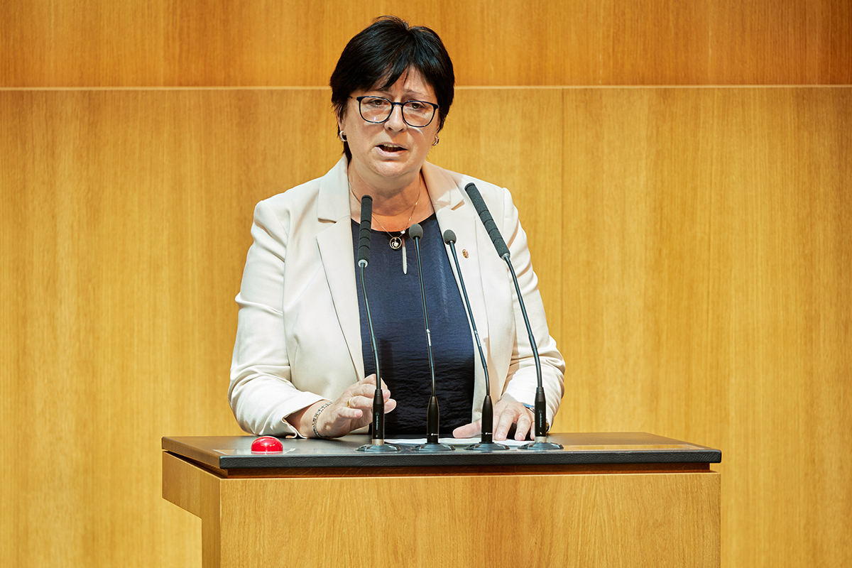 FPÖ Milletvekili Rosa Ecker, üzerinde siyah bir ceket ve kırmızı bir bluzla Avusturya Ulusal Meclisi (Nationalrat) kürsüsünde mikrofon başında konuşma yapıyor. Arka planda meclis salonunun ahşap detayları ve meclis başkanlığı kürsüsü görülüyor.
