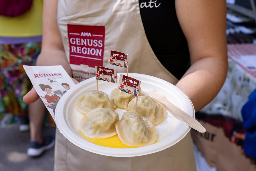 Elde tutulan beyaz tabakta kürdanla servis edilen beş adet yerel mantı benzeri tadımlık ürün. Üzerinde küçük festival bayrakları bulunuyor.