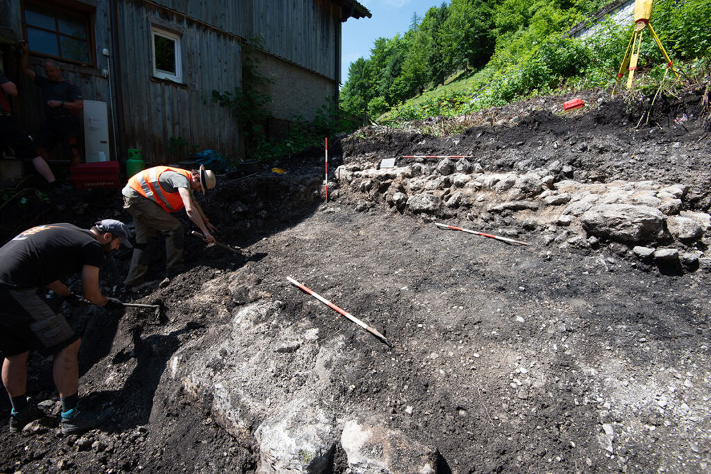 Hallstatt'taki Riezinger-Ausgrabung sahasında, iki arkeoloğun kazı yaparken ve ölçüm çubukları kullanırken çekilmiş fotoğrafı. Arka planda ahşap bina ve ormanlık alan görülüyor.