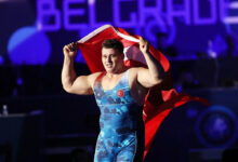 Milli güreşçi Rıza Kayaalp, mavi mayosuyla minder üzerinde sırtına aldığı Türk bayrağını iki eliyle tutarak zafer turu atıyor. Arka planda mor ışıklı bir arena atmosferi görülüyor.