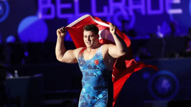 Milli güreşçi Rıza Kayaalp, mavi mayosuyla minder üzerinde sırtına aldığı Türk bayrağını iki eliyle tutarak zafer turu atıyor. Arka planda mor ışıklı bir arena atmosferi görülüyor.