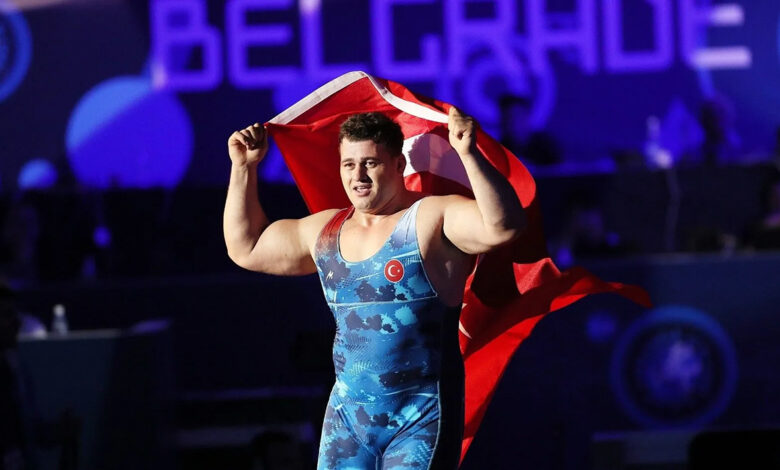 Milli güreşçi Rıza Kayaalp, mavi mayosuyla minder üzerinde sırtına aldığı Türk bayrağını iki eliyle tutarak zafer turu atıyor. Arka planda mor ışıklı bir arena atmosferi görülüyor.