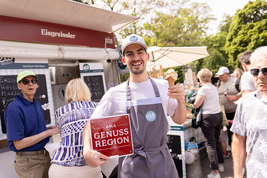Elinde külah dondurma ve kırmızı AMA Genuss Region levhası tutan güler yüzlü bir festival katılımcısı.