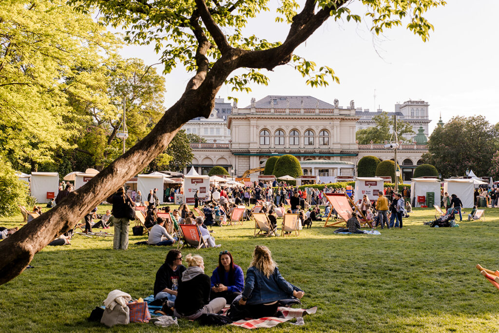 Viyana Stadtpark'ta güneşli bir günde festival alanında çimlere yayılmış insanlar, dinlenme koltukları ve arkada tarihi Kursalon binası.