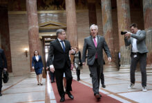 Avusturya Ulusal Meclis Başkanı Walter Rosenkranz ve Moldova Parlamento Başkanı Igor Grosu, Viyana'daki parlamento binasının görkemli sütunlu salonunda kırmızı halı üzerinde yürürken sohbet ediyor. Arka planda devasa mermer sütunlar ve bir fotoğrafçı görülüyor.