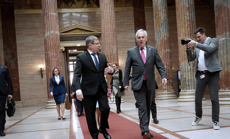 Avusturya Ulusal Meclis Başkanı Walter Rosenkranz ve Moldova Parlamento Başkanı Igor Grosu, Viyana'daki parlamento binasının görkemli sütunlu salonunda kırmızı halı üzerinde yürürken sohbet ediyor. Arka planda devasa mermer sütunlar ve bir fotoğrafçı görülüyor.