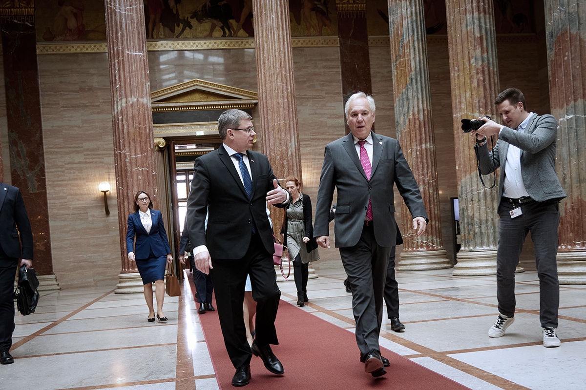 Avusturya Ulusal Meclis Başkanı Walter Rosenkranz ve Moldova Parlamento Başkanı Igor Grosu, Viyana'daki parlamento binasının görkemli sütunlu salonunda kırmızı halı üzerinde yürürken sohbet ediyor. Arka planda devasa mermer sütunlar ve bir fotoğrafçı görülüyor.