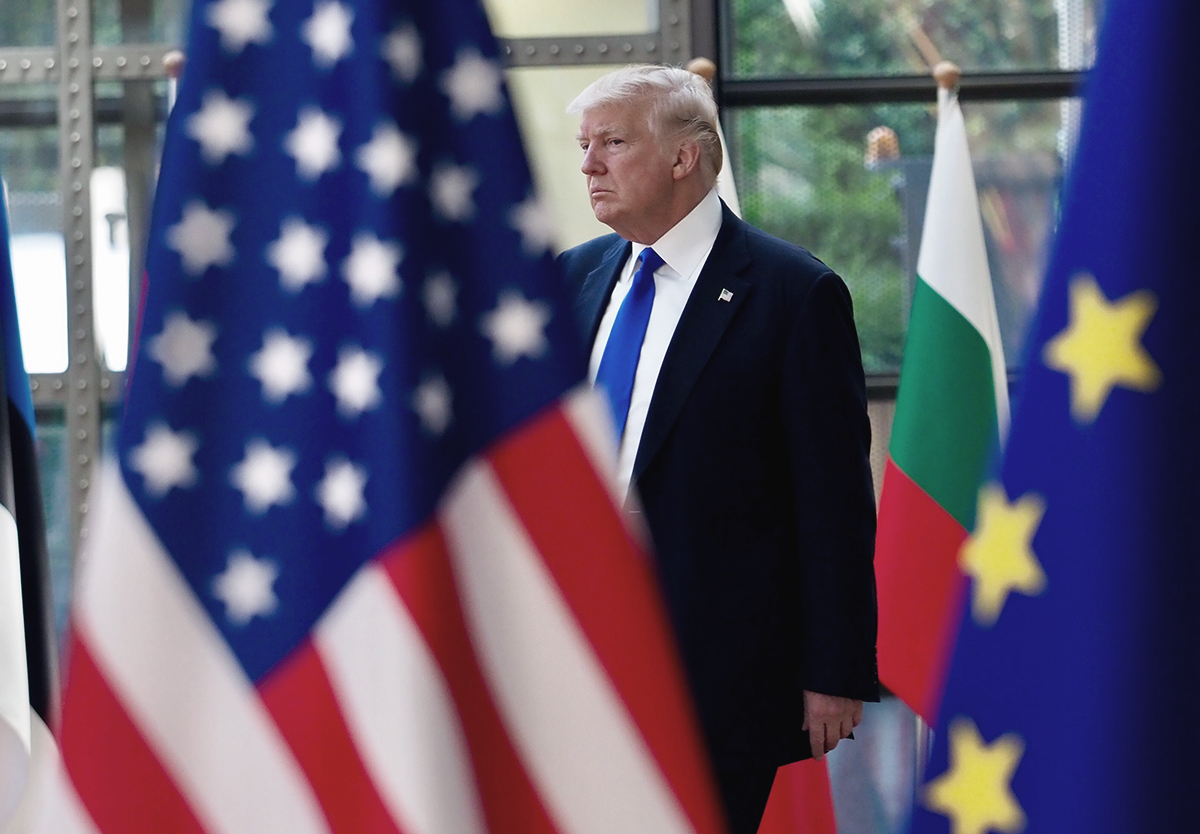 Donald Trump AB ve ABD bayrakları arasından geçerken transatlantik ittifakındaki çatlağı simgeleyen görsel.