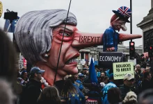 Londra'da Brexit karşıtı protestoda, burnu uzamış hicivli Theresa May ve mini Boris Johnson karikatürleri maketi. Üzerinde 'BREXIT' ve 'ECONOMY' yazılı bavul var.