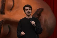 Komedyen Deniz Göktaş Ölü Deniz stand-up gösterisinde elinde mikrofonla sahnede konuşuyor