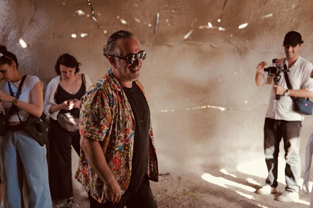 GORA 4 GORA filminin mağara benzeri setinde Cem Yılmaz renkli gömleği ve güneş gözlüğüyle teknik ekibin önünde gülümsüyor.