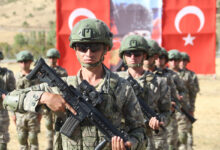 Kamuflaj üniformalı, kasklı ve güneş gözlüklü Türk askerleri ellerinde piyade tüfekleriyle, arka plandaki büyük bir Türk bayrağının önünde sıra halinde duruyor.