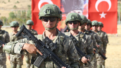 Kamuflaj üniformalı, kasklı ve güneş gözlüklü Türk askerleri ellerinde piyade tüfekleriyle, arka plandaki büyük bir Türk bayrağının önünde sıra halinde duruyor.
