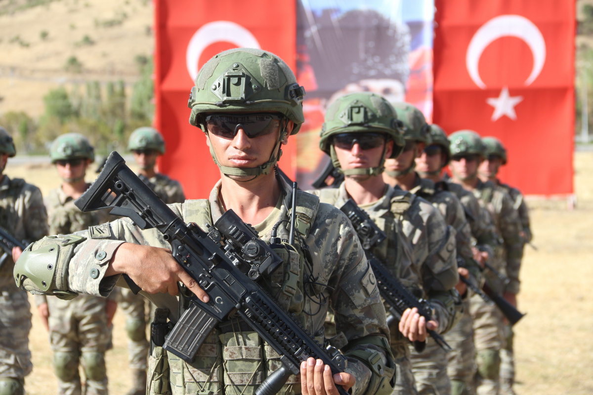 Kamuflaj üniformalı, kasklı ve güneş gözlüklü Türk askerleri ellerinde piyade tüfekleriyle, arka plandaki büyük bir Türk bayrağının önünde sıra halinde duruyor.
