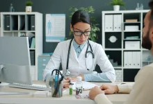 Muayenehanede masada tıbbi geçmiş belgesini düzenleyen doktor ve karşısında oturan hasta.