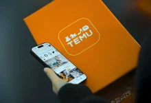 Bir kişinin elinde Temu alışveriş uygulamasının açık olduğu akıllı telefon ve arka planda ikonik turuncu renkli Temu kargo paketi kutusu
