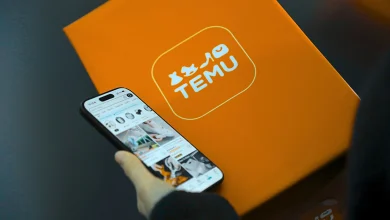 Bir kişinin elinde Temu alışveriş uygulamasının açık olduğu akıllı telefon ve arka planda ikonik turuncu renkli Temu kargo paketi kutusu