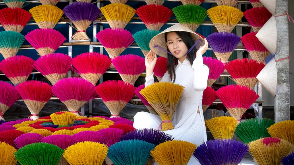 Vietnam geleneksel Ao Dai elbisesi ve konik şapkasıyla çok renkli tütsü duvarı önünde sarı tütsü demeti tutan gülümseyen kadın.