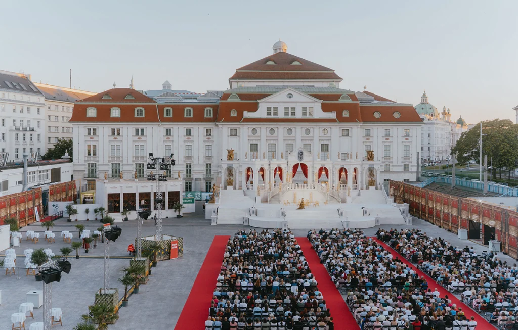 Viyana Opera Yazı festivalinin Heumarkt'ta kurulan geniş açık hava arenası, kırmızı halı detayı, koltukları dolduran yüzlerce sanatsever ve görkemli beyaz sahne tasarımı.