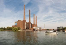 Volkswagen Wolfsburg fabrikası dev bacaları ve bina üzerindeki VW logosu, şirketin geleceği risk altında haber görseli.