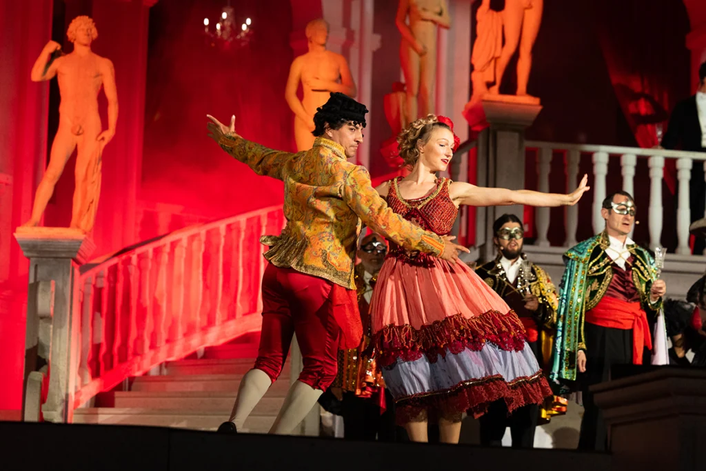 Viyana Opera Yazı festivalinde Heumarkt açık hava arenasındaki görkemli sahnede, kırmızı ışıklar altında dans eden opera sanatçıları ve arkada maskeli koro üyeleri.