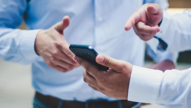 Mavi ve beyaz gömlekli iki kişinin yakın planda bir akıllı telefonu tutarken ve parmakla cihazı işaret ederken çekilmiş fotoğrafı.