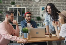 Modern bir ofis ortamında bilgisayar ekranı başında aktif şekilde iletişim kuran ve tartışan çok kültürlü genç profesyoneller grubu.