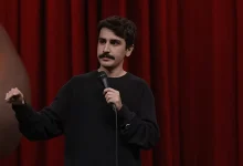 Komedyen Deniz Göktaş, kırmızı sahne perdesi önünde siyah sweatshirtü ile el jesti yaparak stand-up gösterisini gerçekleştiriyor.