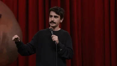 Komedyen Deniz Göktaş, kırmızı sahne perdesi önünde siyah sweatshirtü ile el jesti yaparak stand-up gösterisini gerçekleştiriyor.