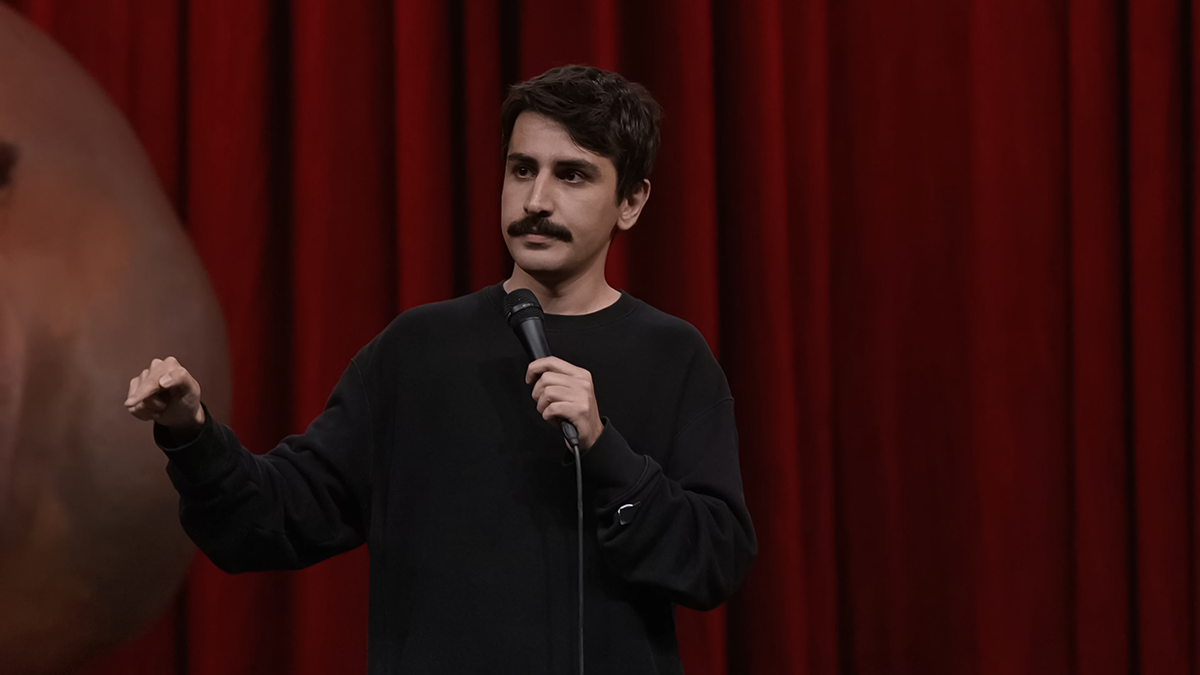 Komedyen Deniz Göktaş, kırmızı sahne perdesi önünde siyah sweatshirtü ile el jesti yaparak stand-up gösterisini gerçekleştiriyor.