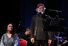 Fuat Saka sahnede mikrofon başında Göç Senfonisi eserini seslendirirken, solist Ioanna Forti ve orkestra sanatçıları sahnede ona eşlik ediyor.