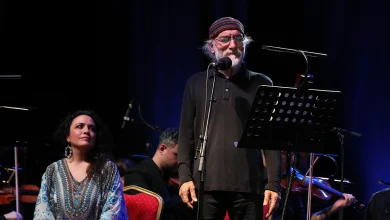 Fuat Saka sahnede mikrofon başında Göç Senfonisi eserini seslendirirken, solist Ioanna Forti ve orkestra sanatçıları sahnede ona eşlik ediyor.