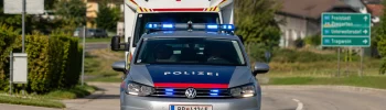 Avusturya'da tepe lambaları açık şekilde yolda ilerleyen gümüş renkli bir polis arabası ve hemen arkasında onu takip eden kırmızı haç logolu bir ambulans.