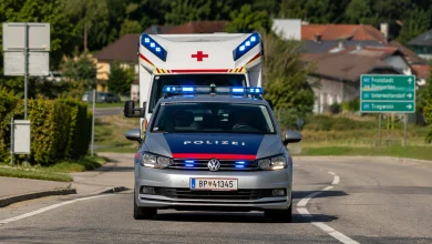 Avusturya'da tepe lambaları açık şekilde yolda ilerleyen gümüş renkli bir polis arabası ve hemen arkasında onu takip eden kırmızı haç logolu bir ambulans.
