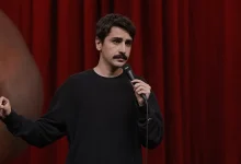 Kırmızı sahne perdesi önünde siyah kazağıyla mikrofon tutarak konuşan komedyen Deniz Göktaş.