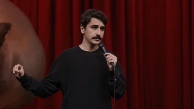 Kırmızı sahne perdesi önünde siyah kazağıyla mikrofon tutarak konuşan komedyen Deniz Göktaş.