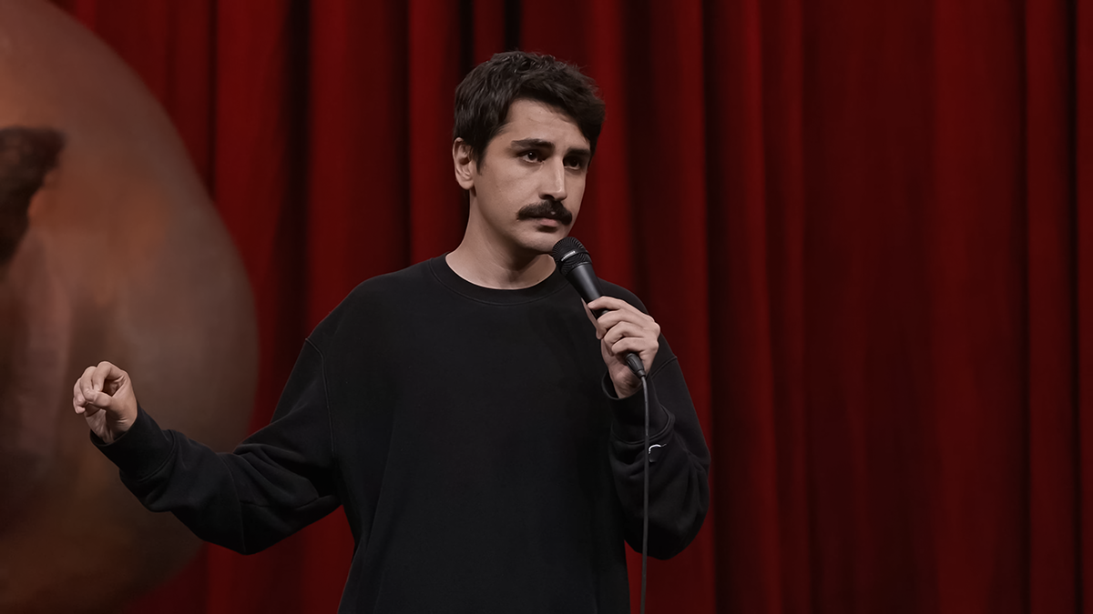 Kırmızı sahne perdesi önünde siyah kazağıyla mikrofon tutarak konuşan komedyen Deniz Göktaş.
