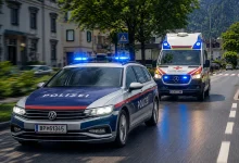 Mavi tepe lambaları açık, hareket halindeki bir Avusturya federal polis arabası ve arkasında bir Kızıl Haç ambulansının yer aldığı şehir içi fotoğrafı.